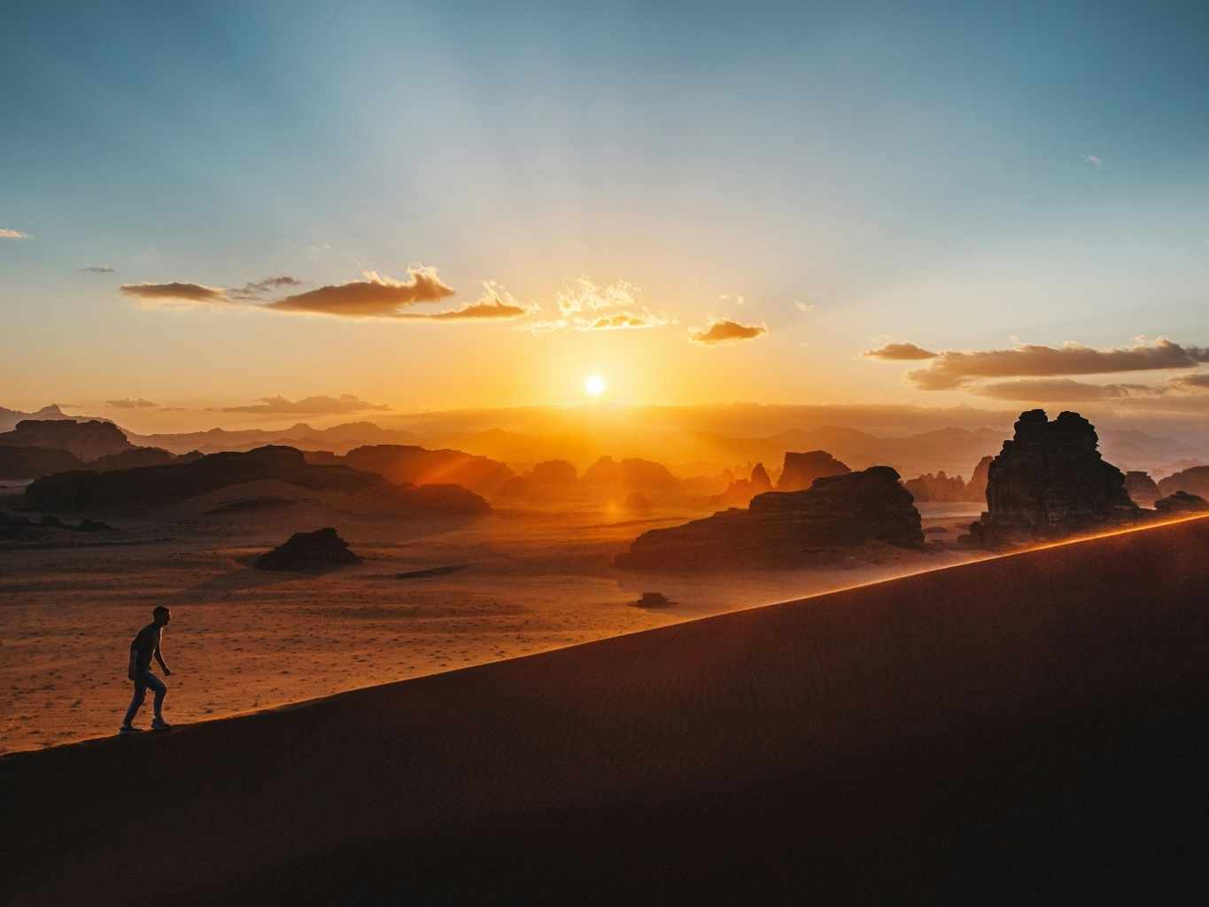 Persona subiendo una duna al amanecer, con el sol elevándose sobre el horizonte en un paisaje desértico